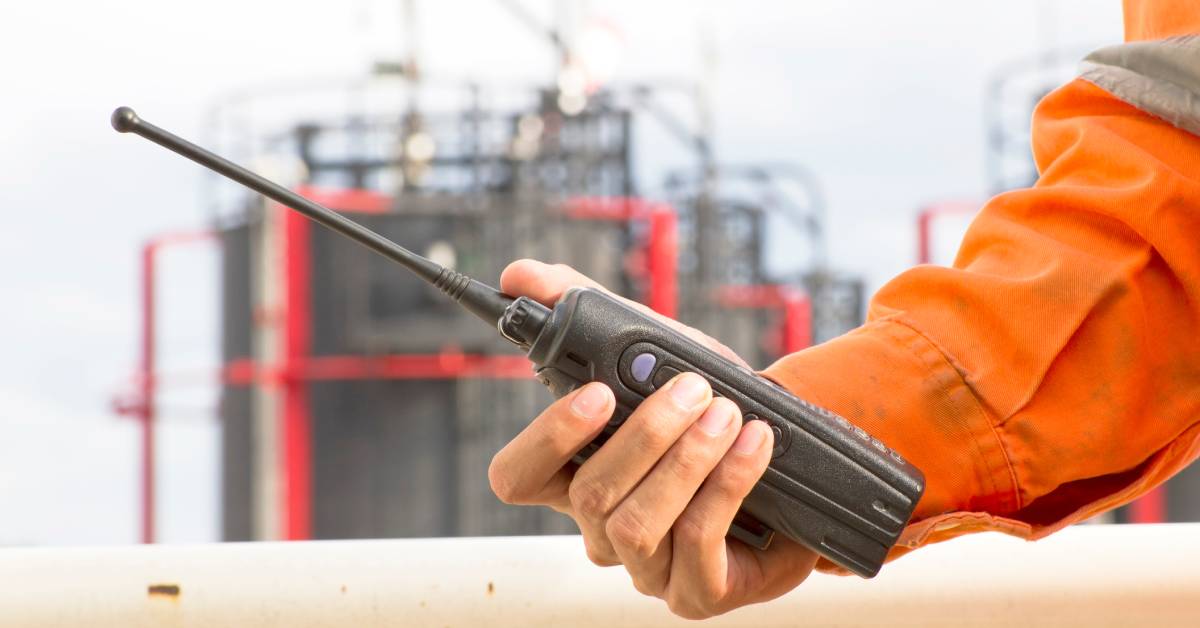 A two-way radio is held in the hand of a male plant worker whose orange protective clothing is dirty.