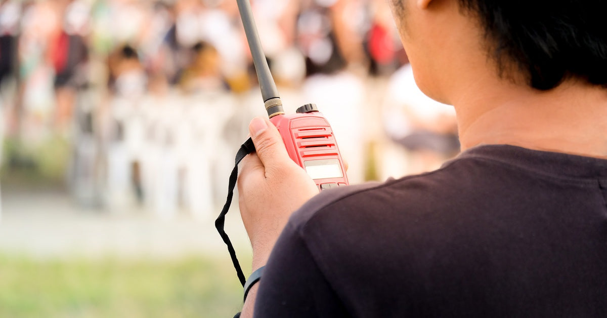 A man wearing a black shirt is holding a red two-way radio to communicate, with a blurry background of an outdoor event.