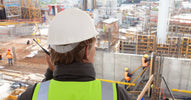 A male supervisor wearing a hardhat is listening to a two-way radio while overseeing a large construction site below him.