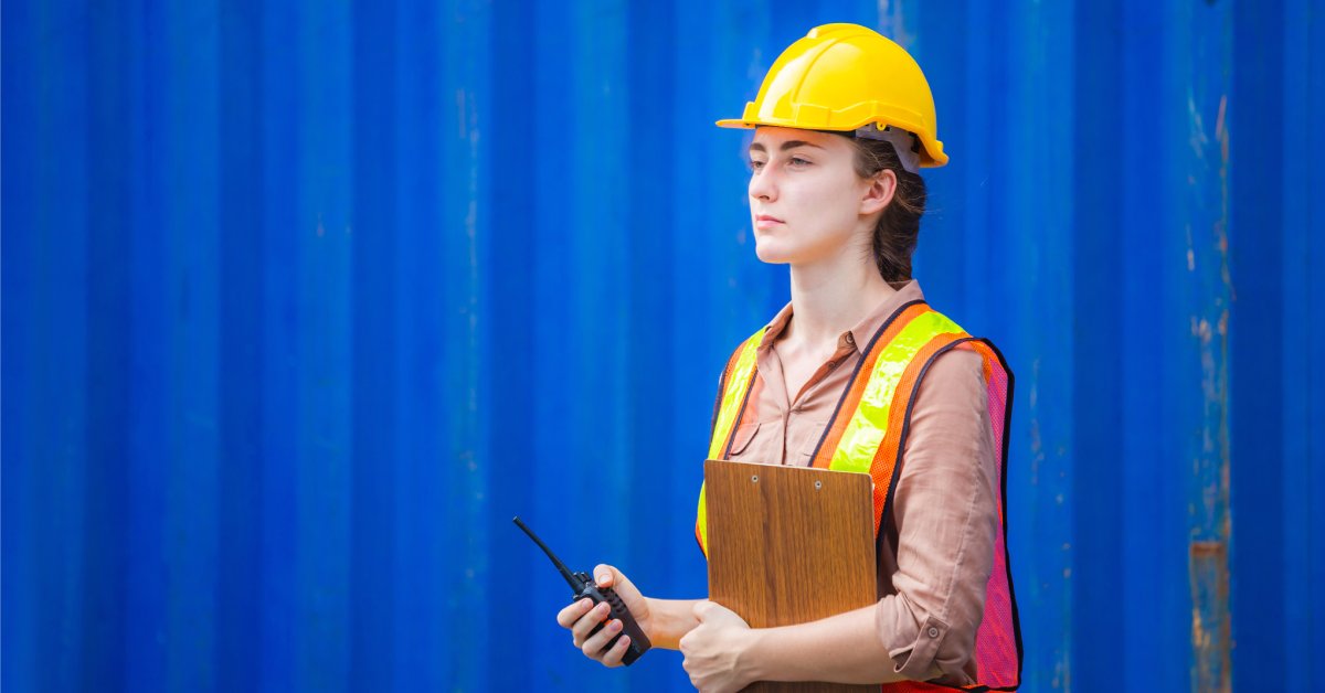 A woman wearing a yellow hard hat and a high-vis safety vest holds a two-way radio and a clipboard.